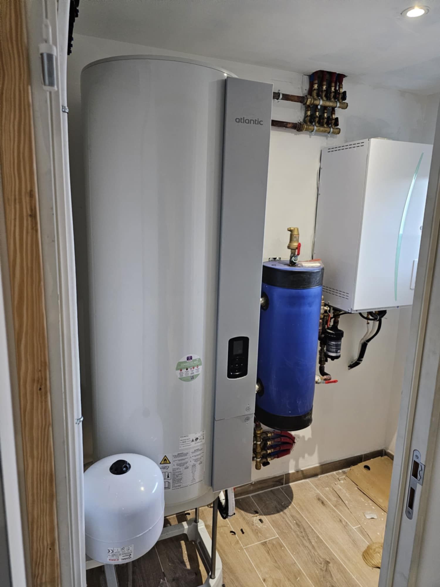 Dans une salle de bains moderne et lumineuse, équipée de deux lavabos et de plantes, un chauffe-eau cylindrique blanc se dresse à côté d'une petite armoire.