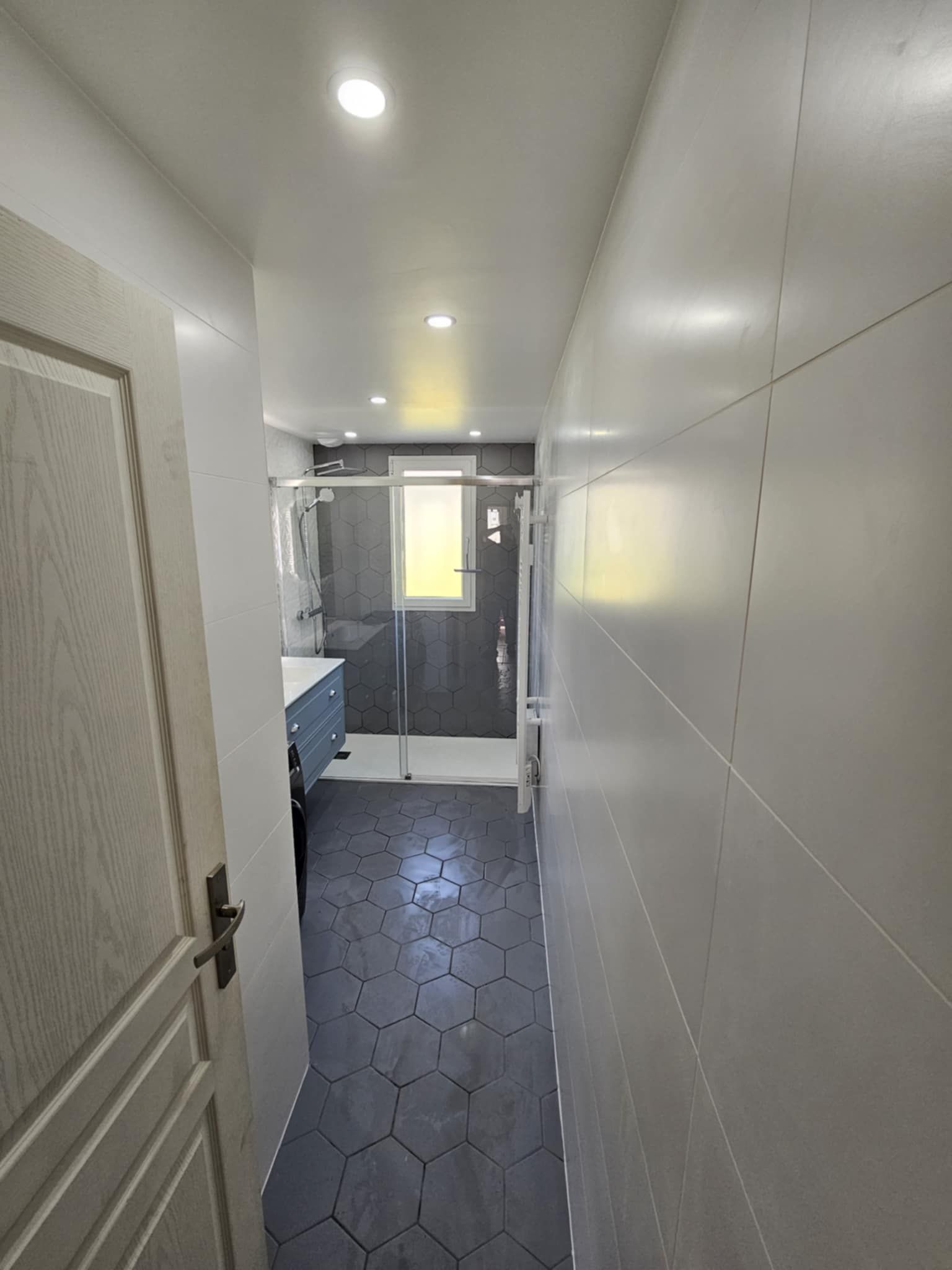 Une personne portant des gants de travail utilise une clé pour réparer les tuyaux métalliques situés sous un lavabo, contre un mur carrelé blanc.