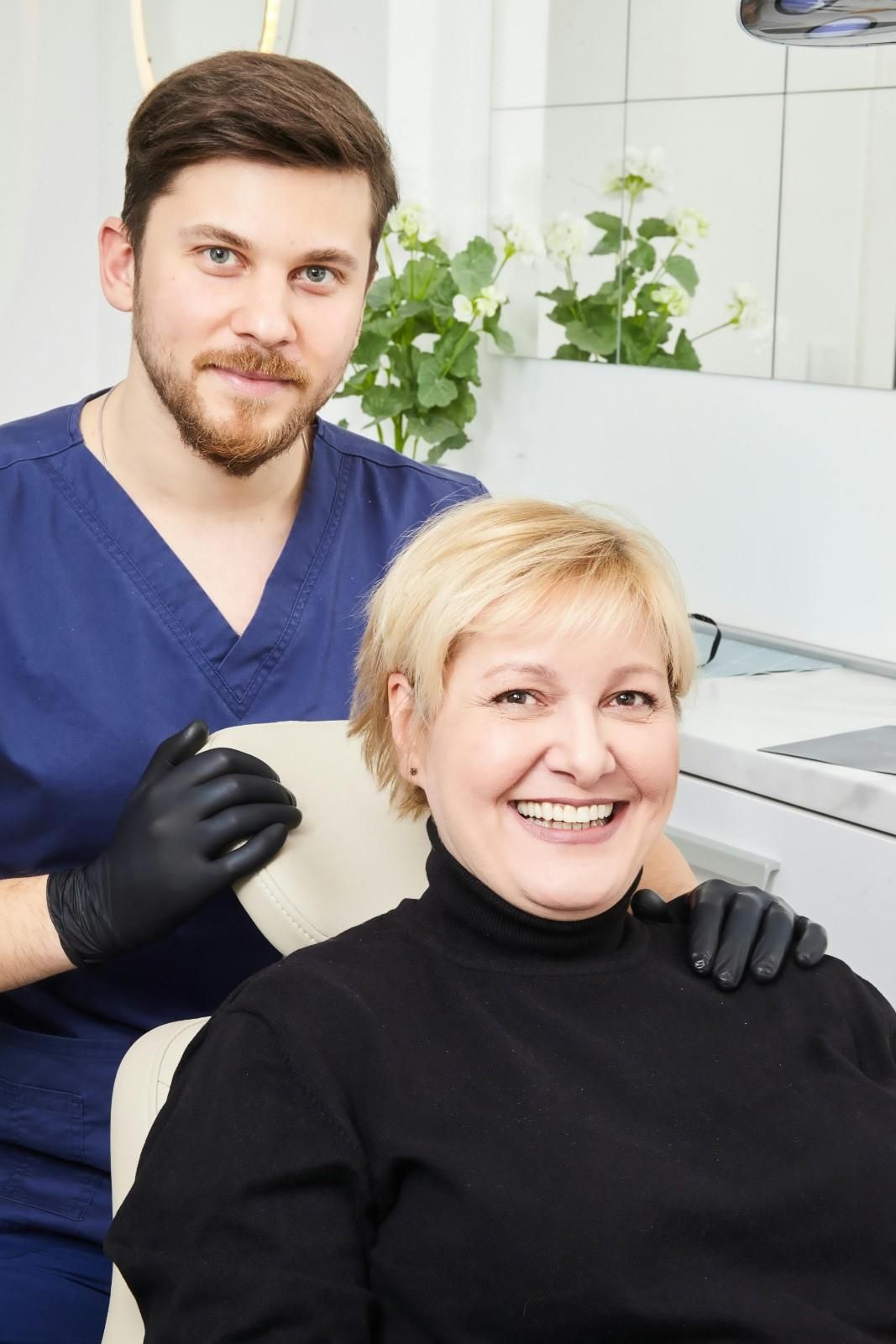 Dentista con bata azul y guantes negros sonriendo junto a un paciente en el sillón dental.