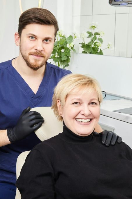 Dentista con bata azul y guantes negros sonriendo junto a un paciente en el sillón dental.