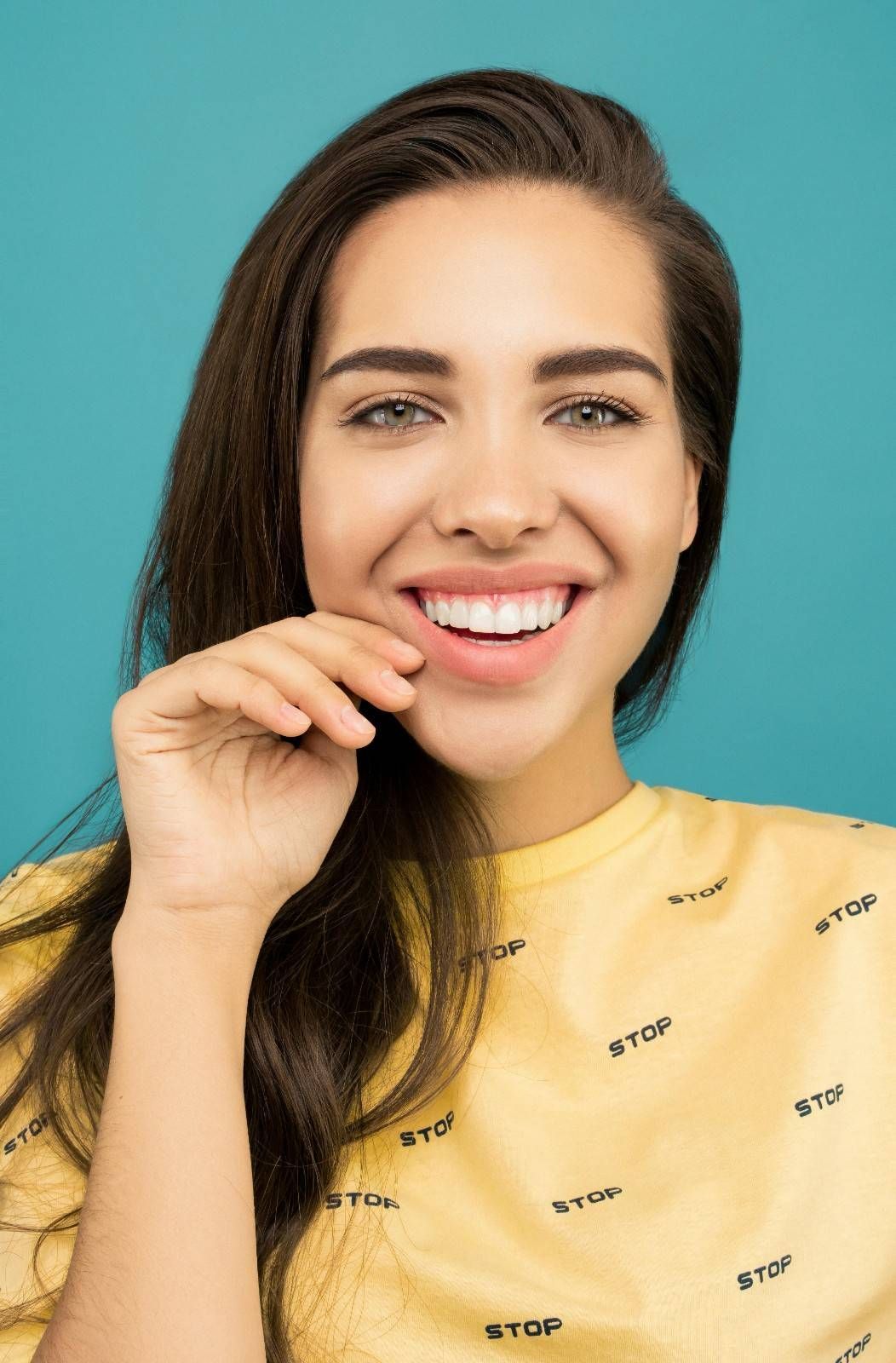 Mujer sonriente con una camisa amarilla sobre un fondo turquesa, con la mano cerca de la boca.