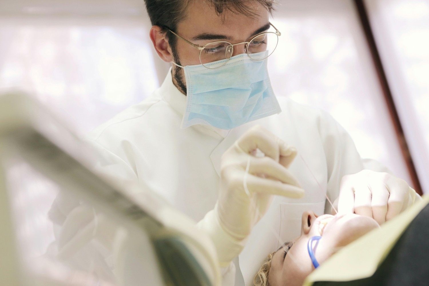 Dentista con mascarilla y guantes atendiendo a un paciente en una clínica bien iluminada.
