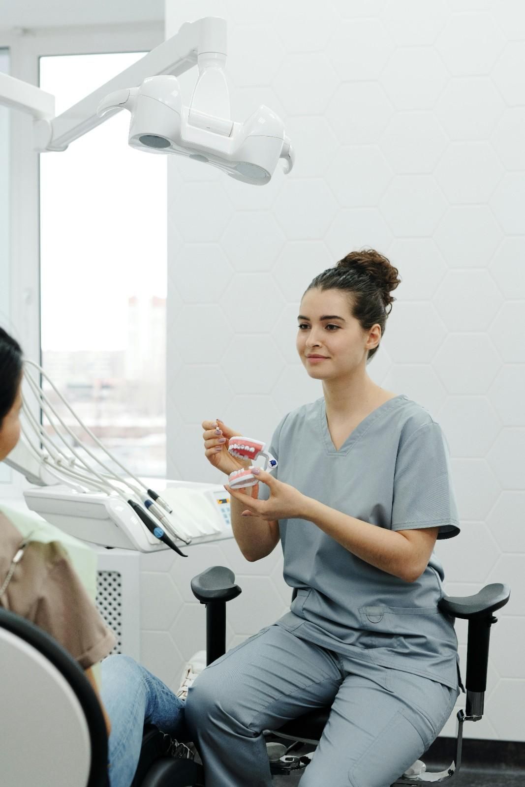 Dentista con bata gris sosteniendo un cepillo de dientes en una sala de examen dental, frente a un paciente sentado.