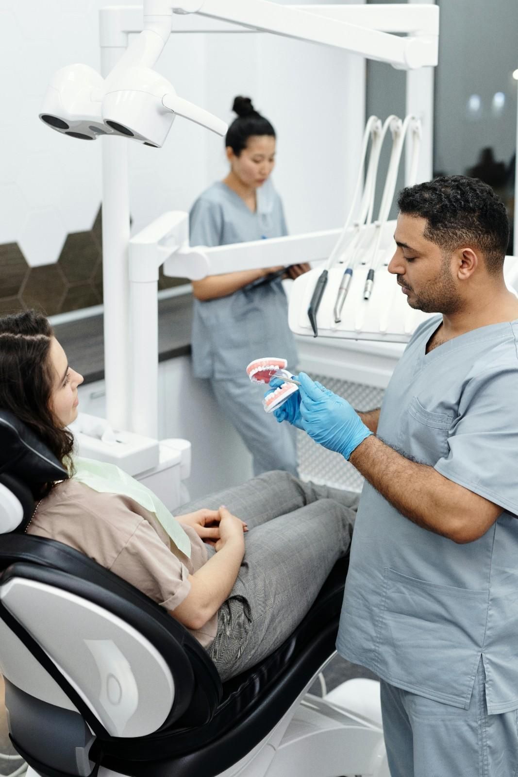 Un dentista se prepara para mostrar un modelo dental a un paciente sentado en una clínica, con su asistente al fondo.