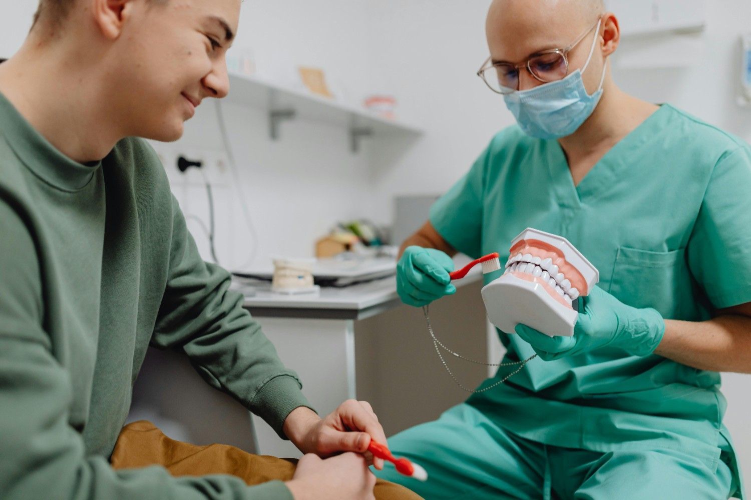 Dentista mostrando un modelo dental a un paciente sonriente en una clínica, ambos con guantes y mascarilla.