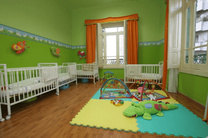 Habitación infantil con cuatro cunas blancas, paredes verdes, cortinas naranjas y tapetes de juego con juguetes sobre suelo de madera.