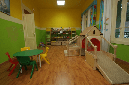 Sala de juegos con una pequeña casa de madera, mesa, sillas y estantes contra paredes amarillas y verdes.