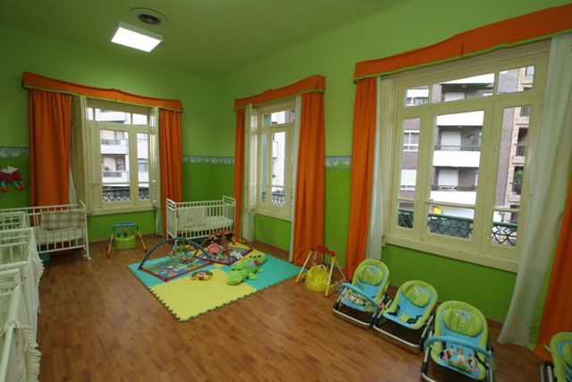 Habitación infantil con suelo de madera, paredes verdes, cortinas naranjas, cunas, juguetes y sillas altas cerca de las ventanas.