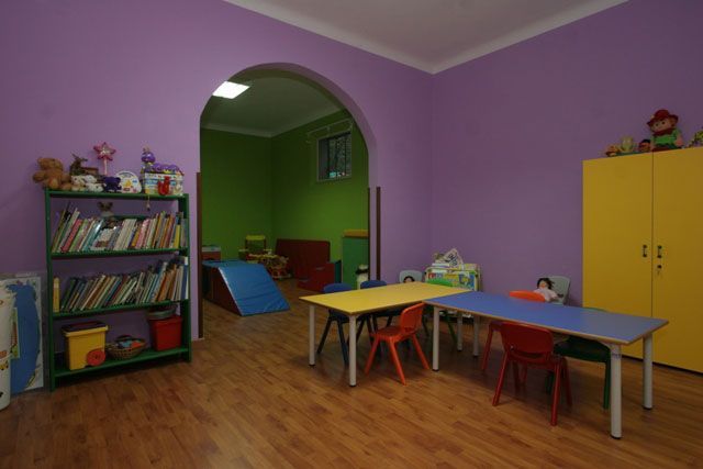 Sala de juegos con paredes moradas, suelo de madera, mesas, sillas y libros.