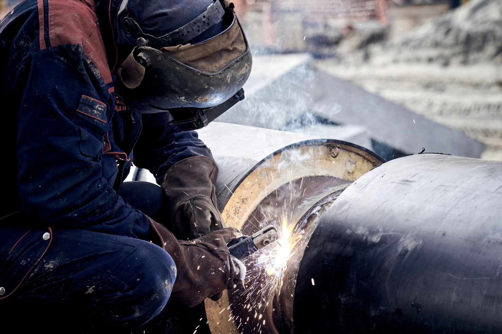 Un soudeur en tenue de protection soude deux gros tuyaux métalliques à l'extérieur ; des étincelles jaillissent.