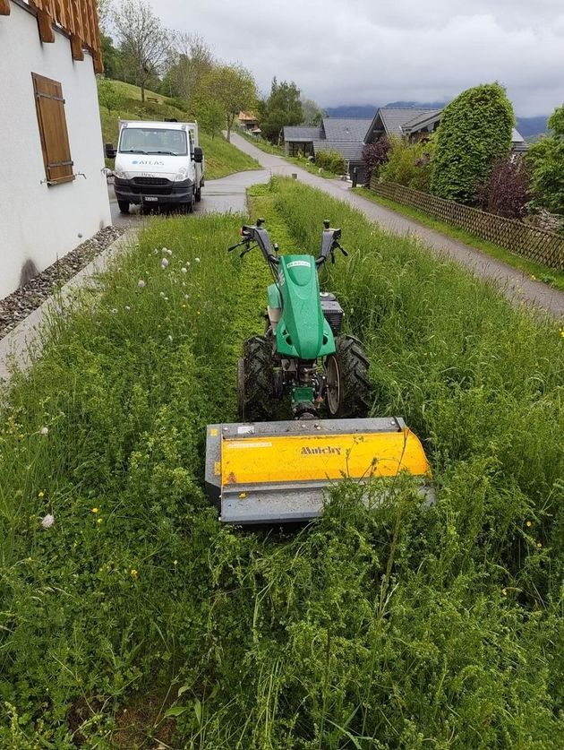 Chemin étroit et humide longeant un terrain vague et des maisons, avec une petite cour clôturée et un ciel couvert.