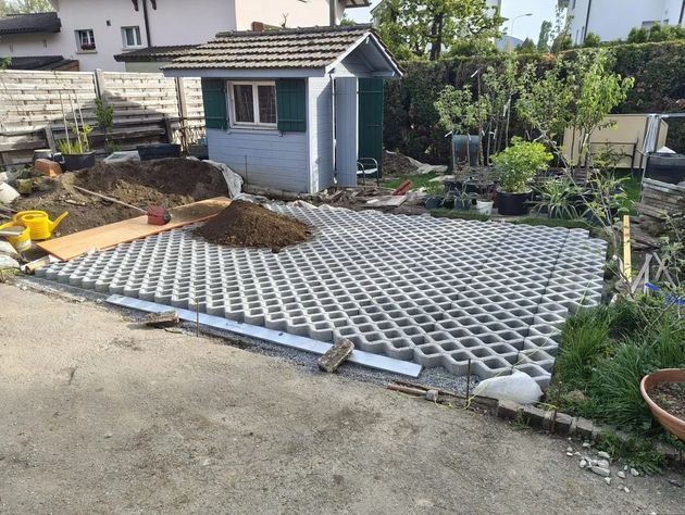 Allée de jardin pavée de dalles grises à treillis, jouxtant un petit abri de jardin bleu et des plantes environnantes.