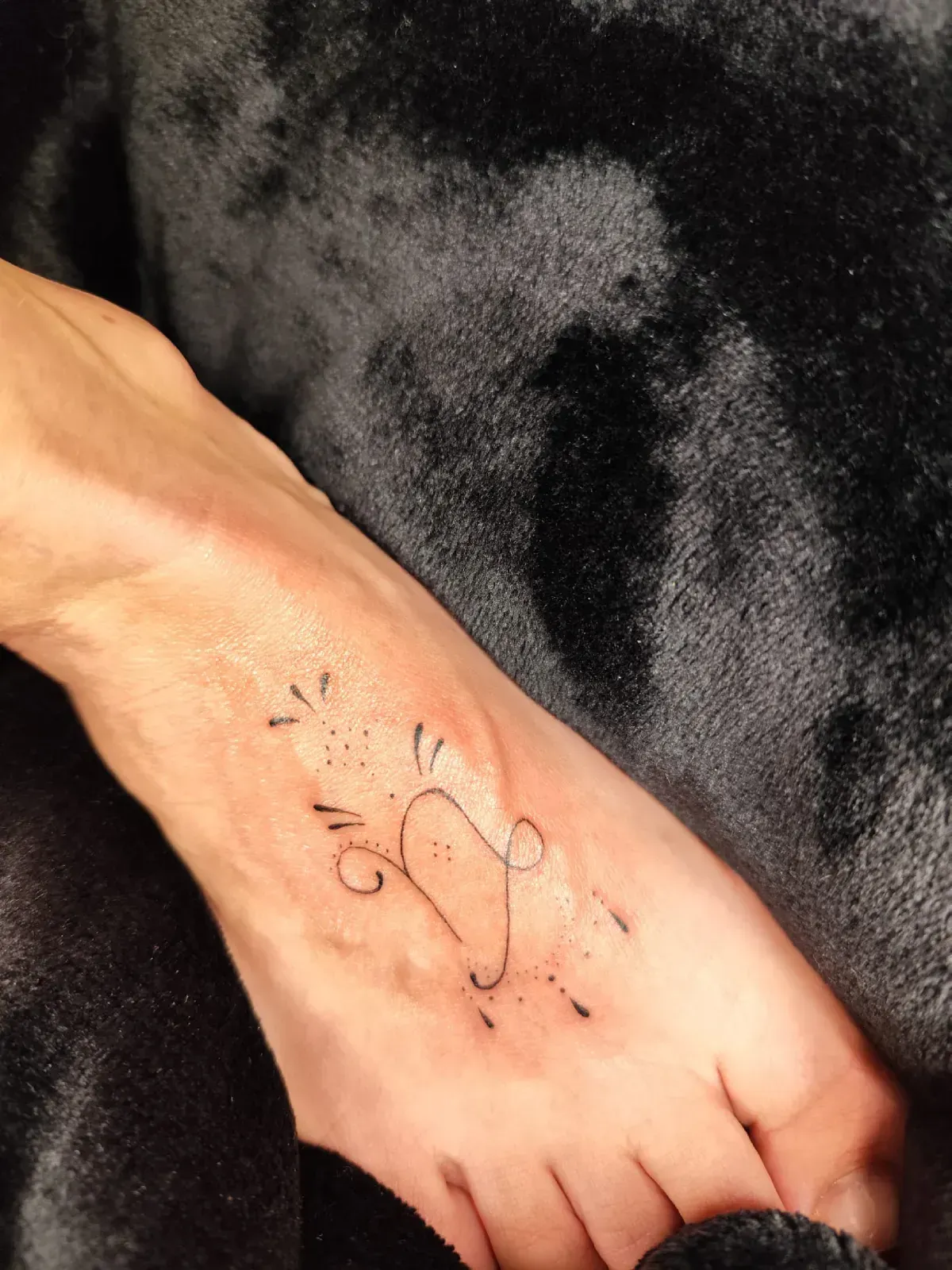 Pie con un delicado tatuaje de tobillera negra y marcas decorativas sobre un fondo de tela oscura.