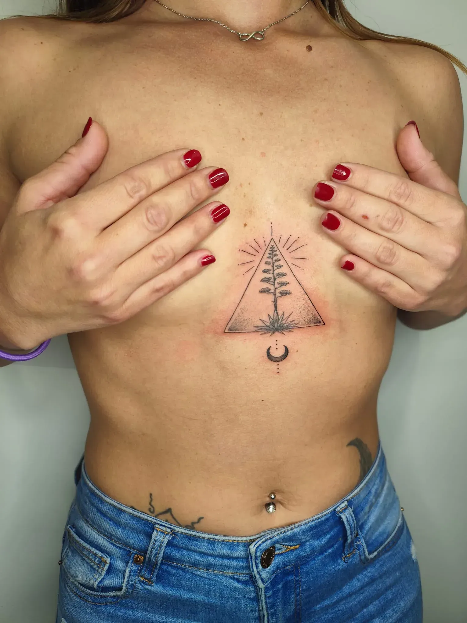 Persona mostrando un tatuaje en el pecho con un símbolo triangular, con las manos sobre el pecho y las uñas pintadas de rojo.
