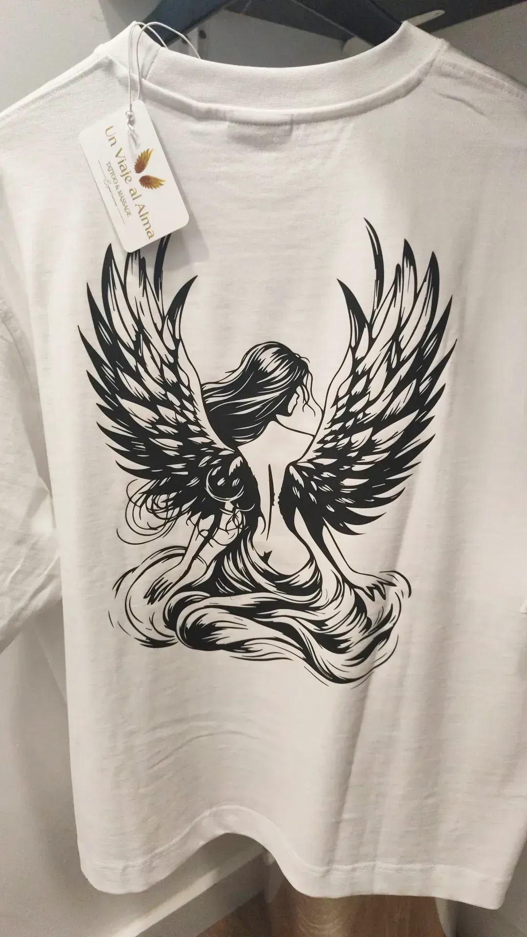 Camiseta blanca con un dibujo de un fénix negro en la espalda, colgada en una percha con etiqueta.