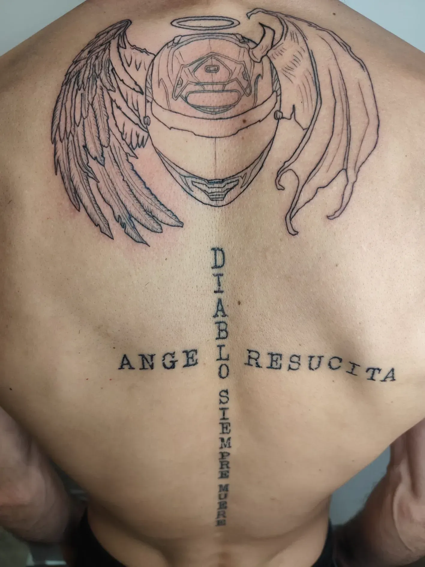 Tatuaje en la espalda con alas de ángel y texto en español “ÁNGEL RESUCITA” y “DIABLO SIENTE”