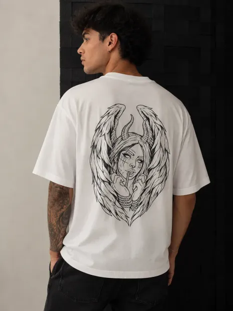 Un hombre con una camiseta blanca estampada está de pie junto a una pared de ladrillos oscuros, mirando hacia un lado.