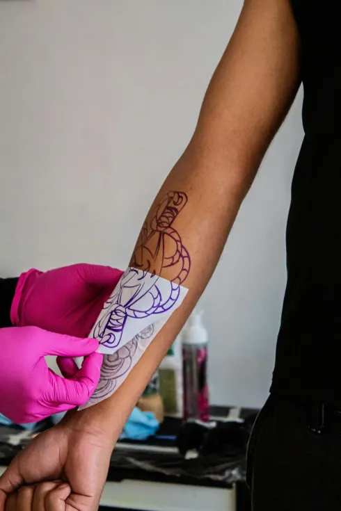 Antebrazo de una persona con una plantilla de tatuaje siendo trazada con tinta morada por manos enguantadas.