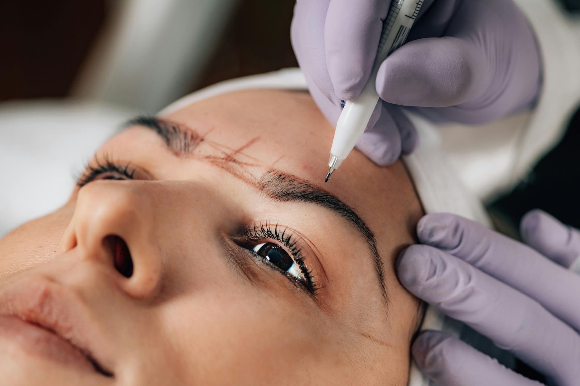 Primer plano de unas manos enguantadas realizando un procedimiento cosmético de cejas en el rostro de una clienta.
