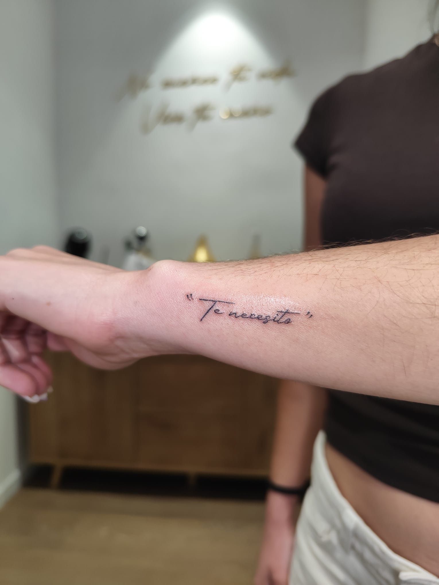 Antebrazo con un pequeño tatuaje en letra cursiva, mostrado en un espacio interior con iluminación tenue.