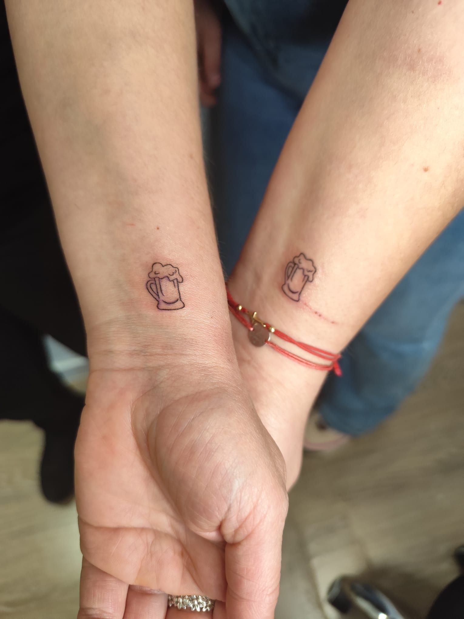 Dos muñecas con pequeños tatuajes a juego y pulseras de hilo rojo, manos unidas frente a un fondo azul.