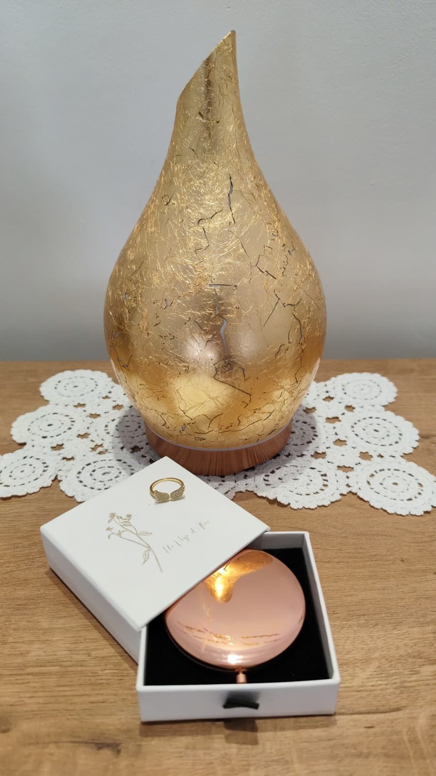 Candelabro dorado en forma de lágrima sobre un tapete de encaje junto a una vela rosa encendida en una caja.