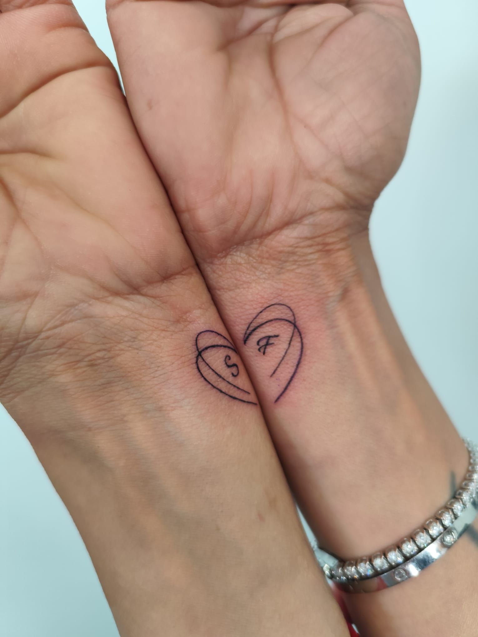 Dos manos entrelazadas, con un pequeño corazón dibujado en la muñeca, cerca de una pulsera de plata.