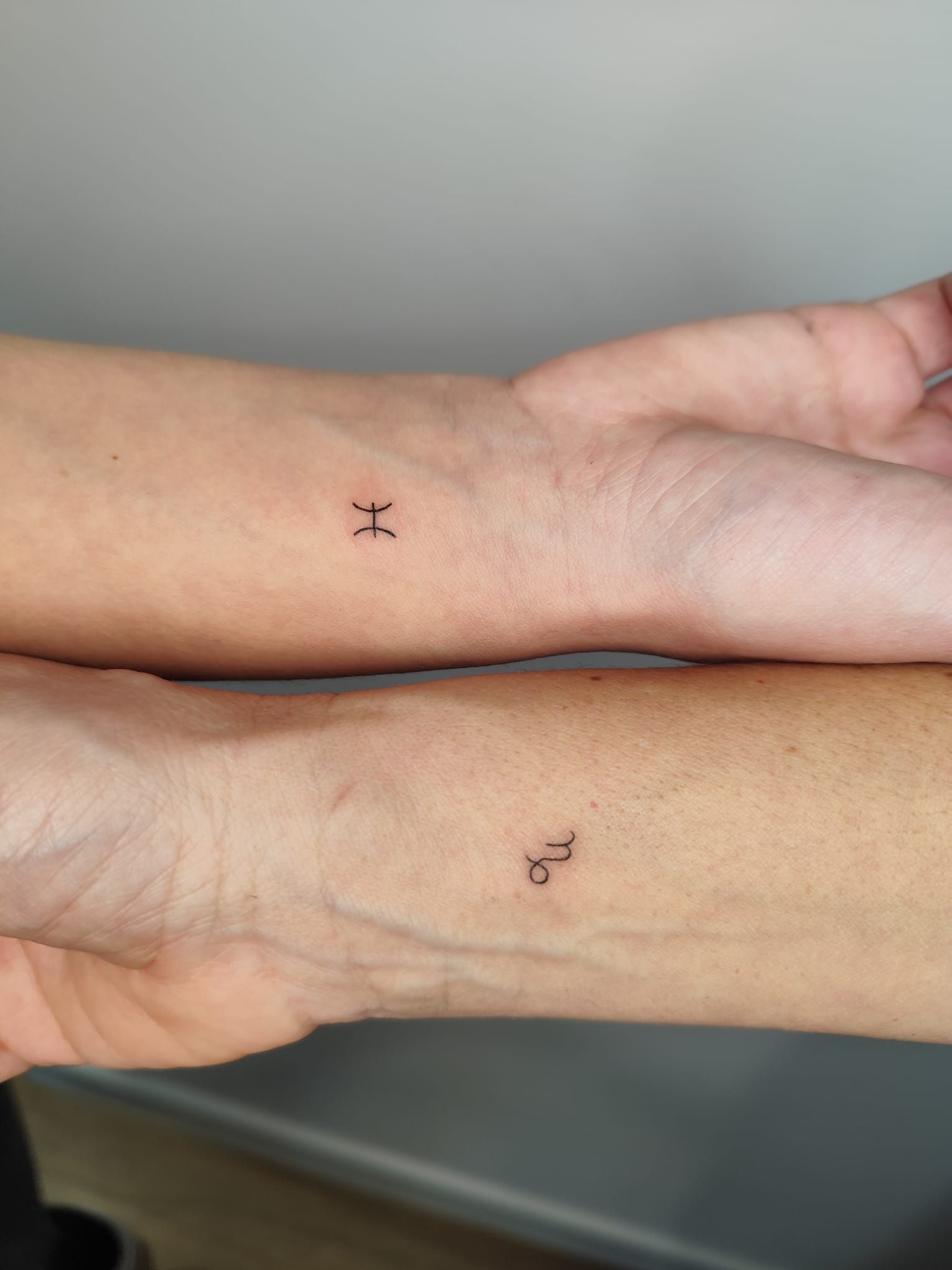 Dos antebrazos uno al lado del otro, cada uno con un pequeño tatuaje negro cerca de la muñeca.