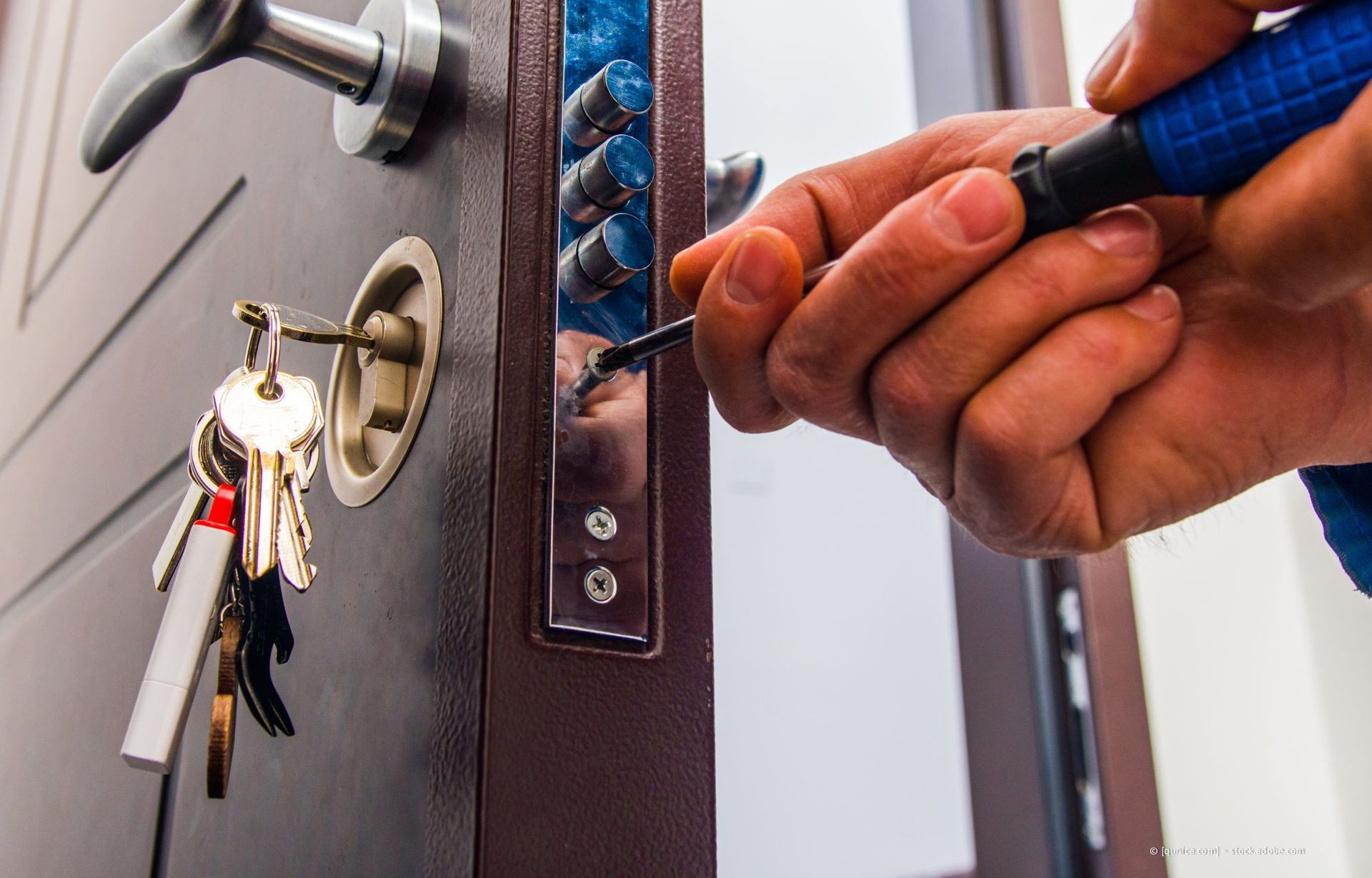 Die Hand einer Person benutzt ein Werkzeug, um ein Schloss zu manipulieren. Schlüssel hängen in der Nähe. Braune Tür, blaues und silbernes Werkzeug.