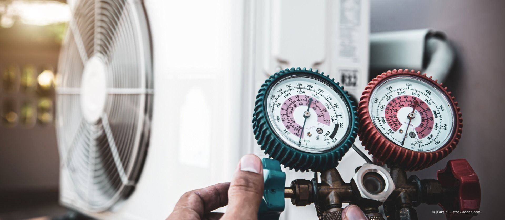 Eine Hand hält zwei Manometer für den HLK-Druck neben einem Ventilator in einem hellen Raum.