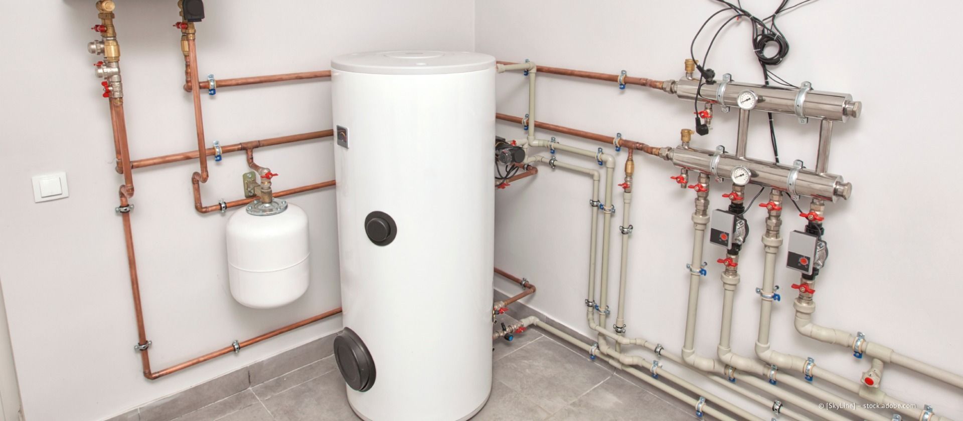 Heizraum mit weißem Warmwasserbereiter, Rohren, Ventilen und einem kleinen Ausdehnungsgefäß an der Wand