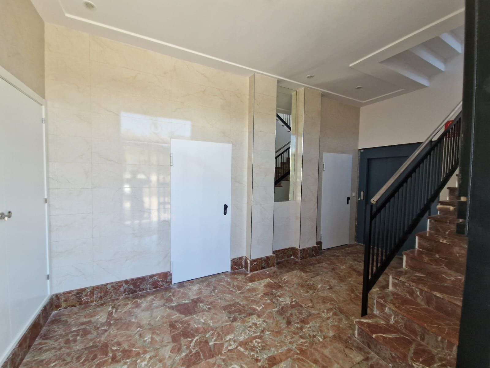 Pasillo con suelo y paredes de mármol, puertas blancas y escalera con barandilla negra.