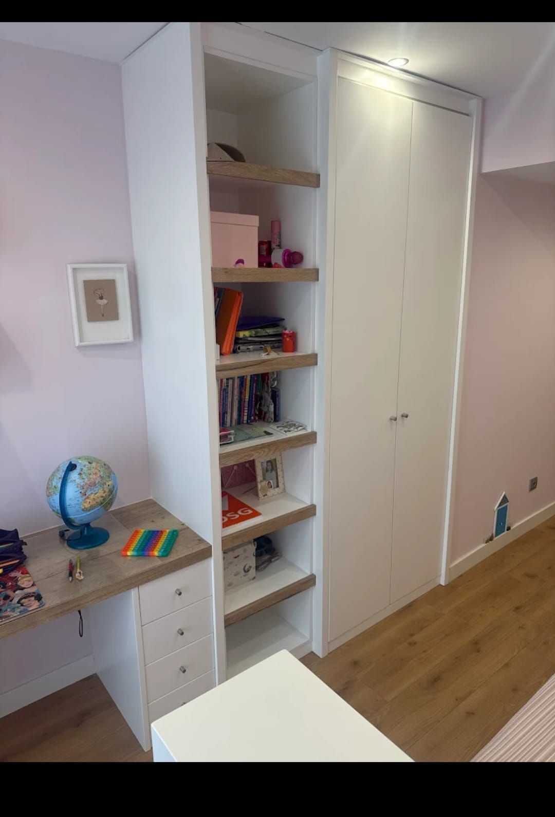 Mueble de almacenaje blanco empotrado con estantes abiertos y armario. Junto a un escritorio en una habitación con paredes rosas.