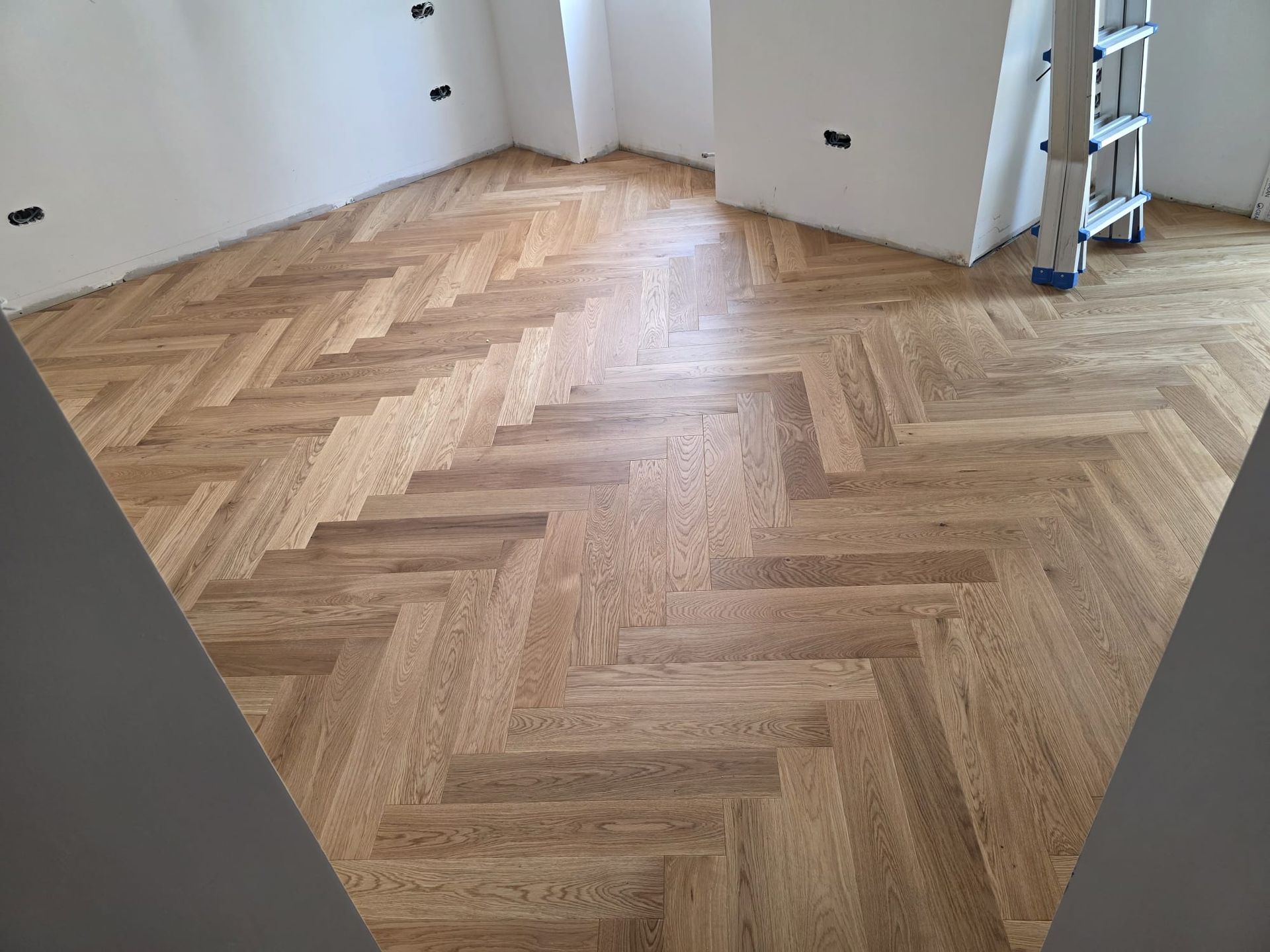Piso de madera con diseño de espiga en una habitación con paredes blancas, sin terminar.