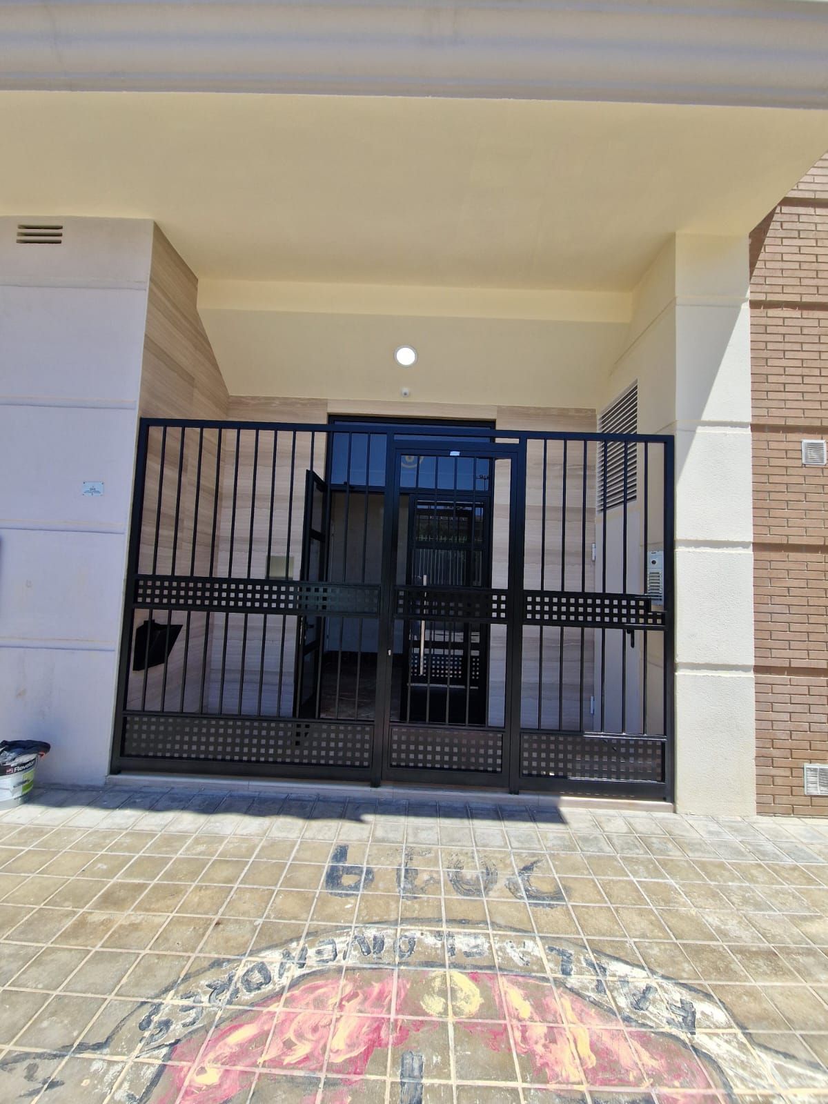 Puerta negra a la entrada del edificio; paredes de color crema y ladrillo, pavimento y puerta azul visible.