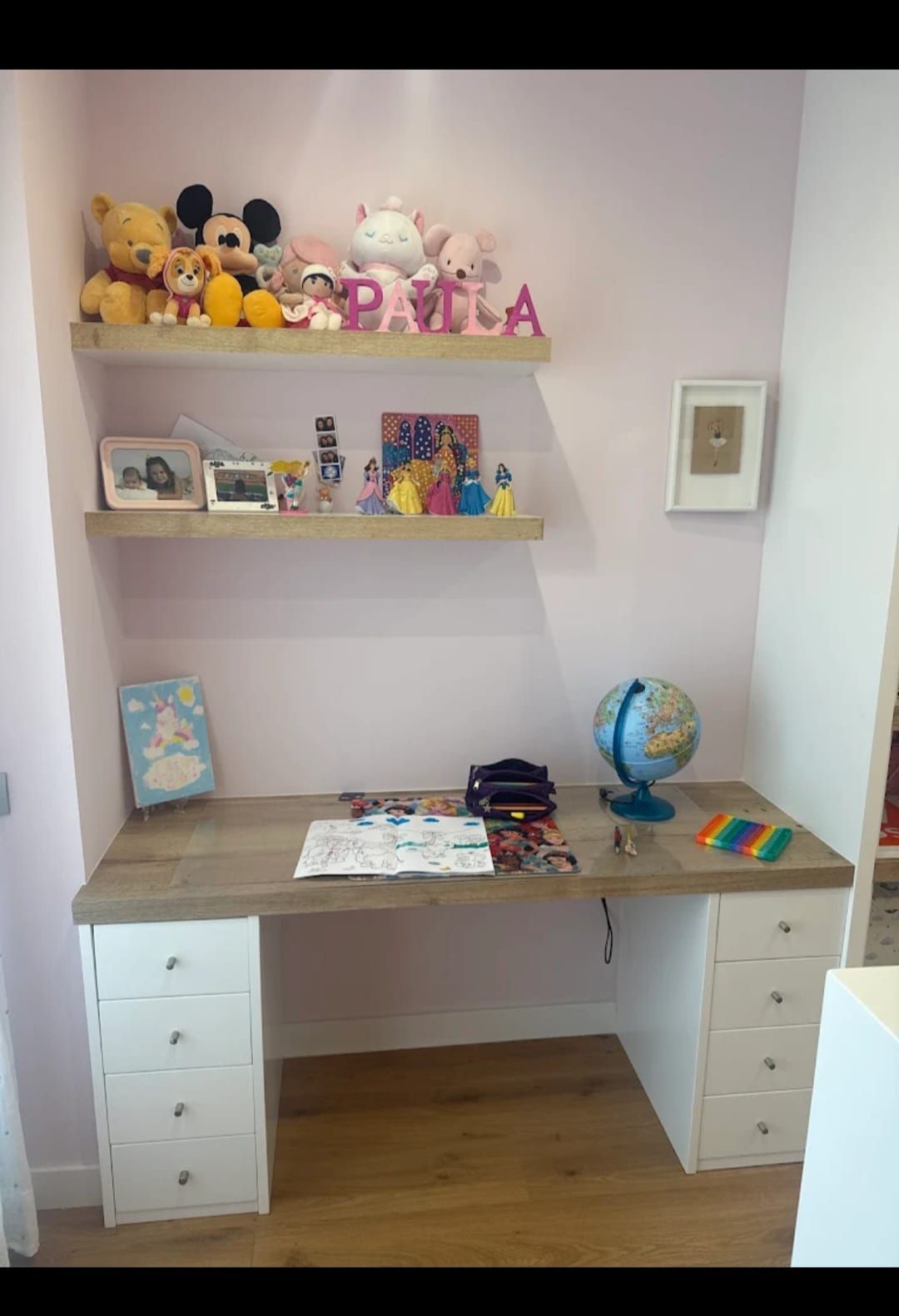 Escritorio infantil con estantes para juguetes y fotos sobre una pared rosa. Tablero de madera y cajones.