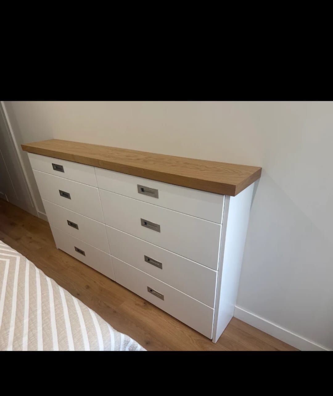 Cómoda blanca con tapa de madera y tiradores plateados sobre una pared blanca y suelo de madera.