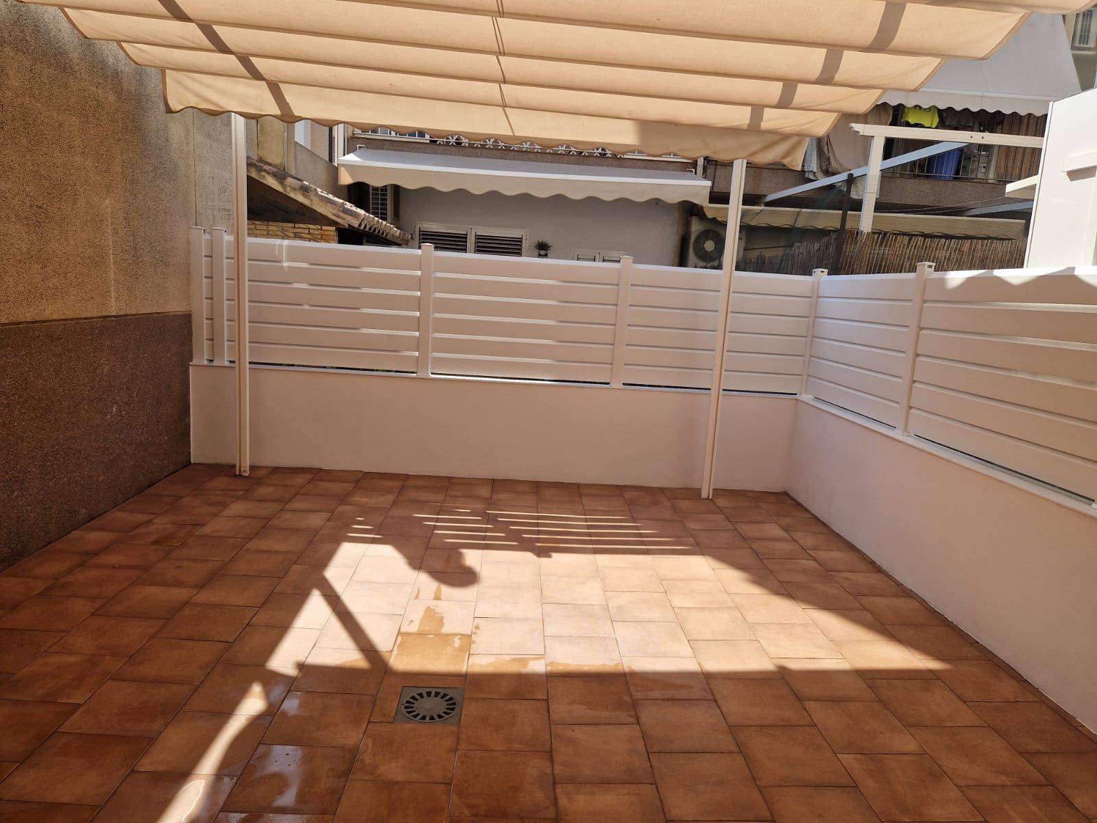 Patio de baldosas con cerca blanca y toldo. La luz del sol crea sombras.