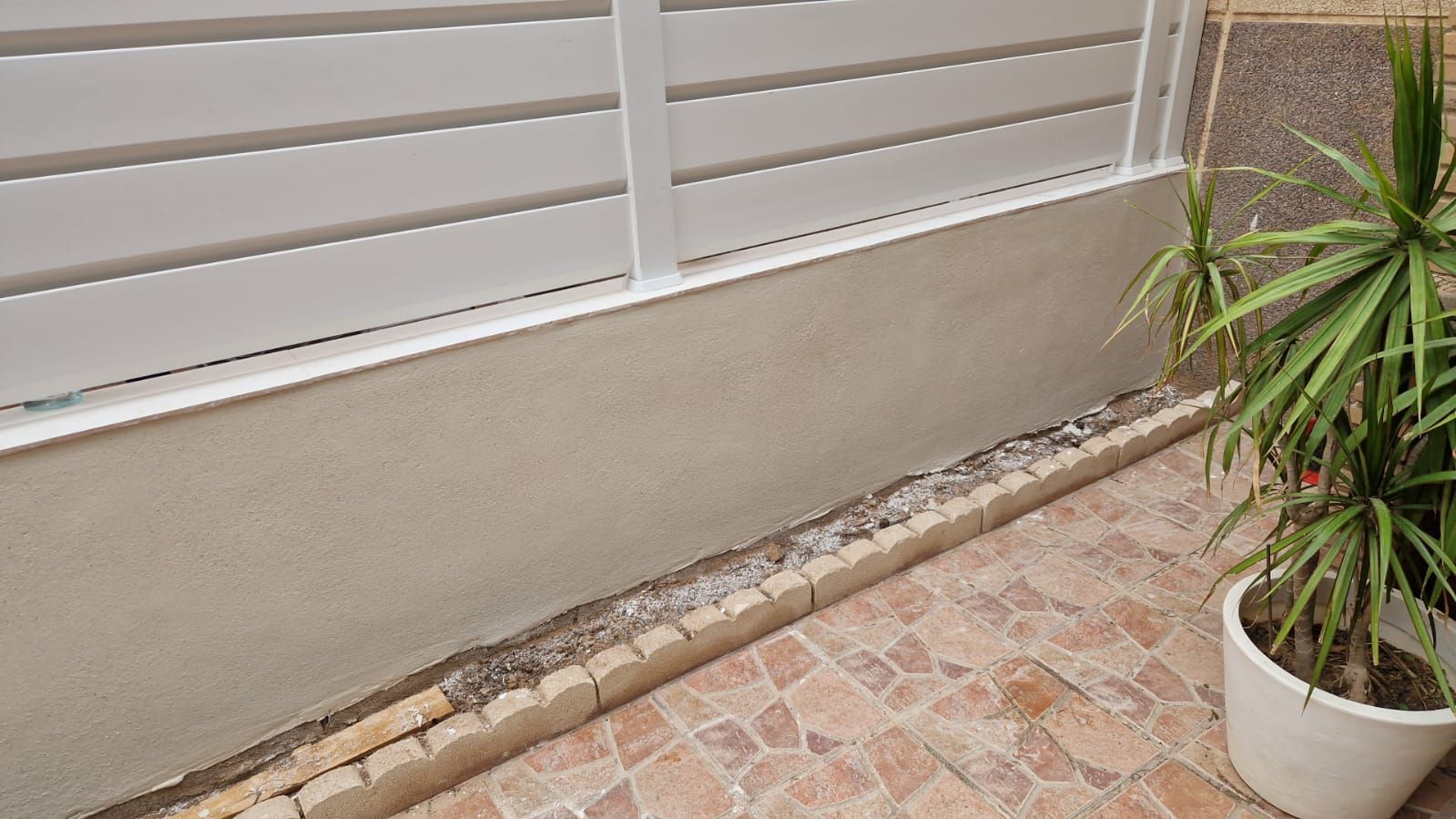 Cerca de listones blancos, pared de estuco, borde de ladrillo y planta en maceta en un patio de baldosas.