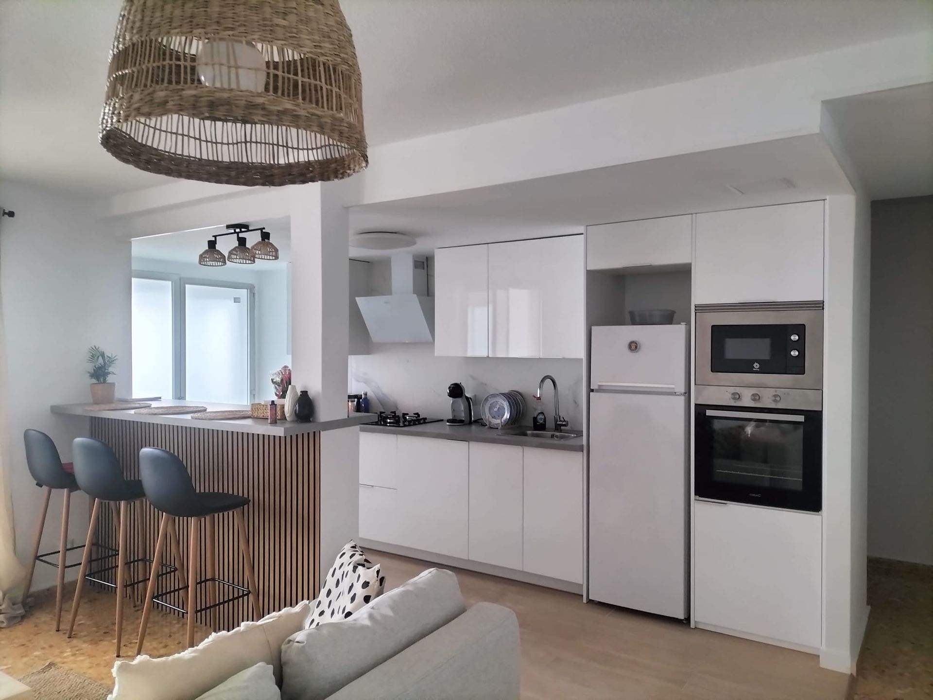 Cocina de concepto abierto con gabinetes blancos, barra de desayuno y lámpara colgante tejida.