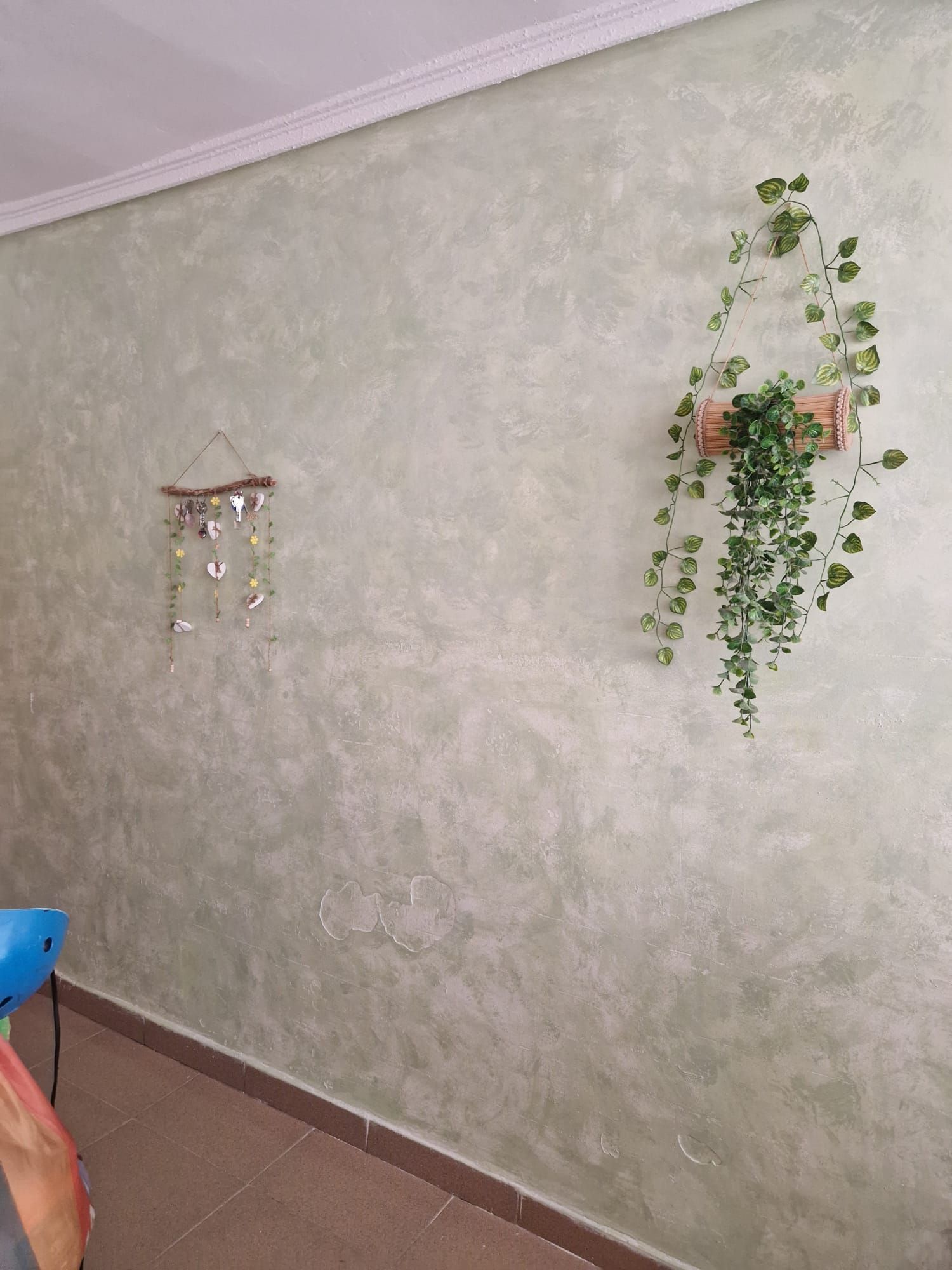 Pared texturizada de color verde pálido con plantas colgantes y un móvil decorativo.