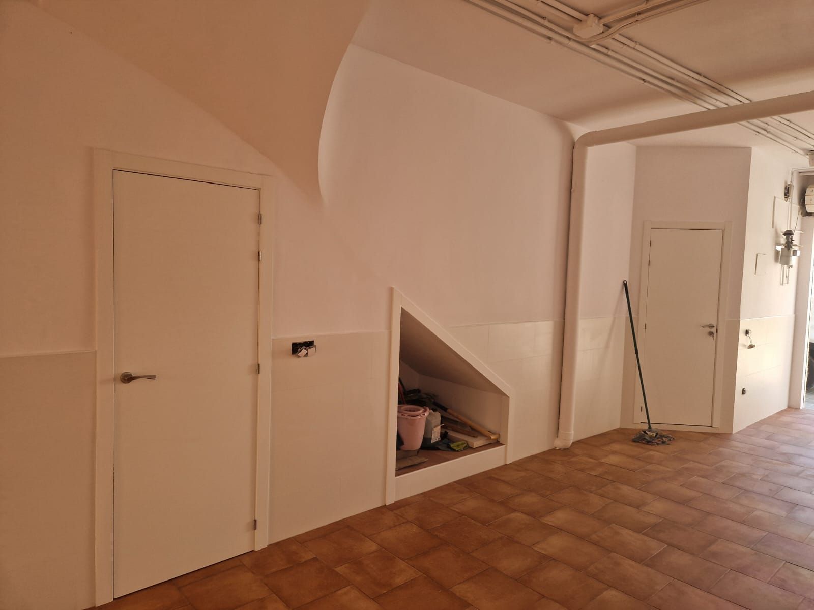 Interior con paredes blancas, dos puertas y un rincón de almacenamiento. Suelo de baldosas marrones.