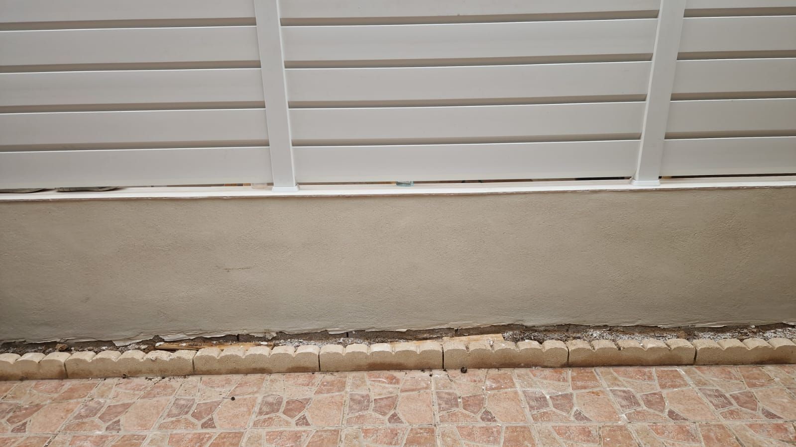 Una valla de listones blancos sobre una pared de estuco gris, encima de una hilera de baldosas de piedra sobre un suelo de baldosas marrones.