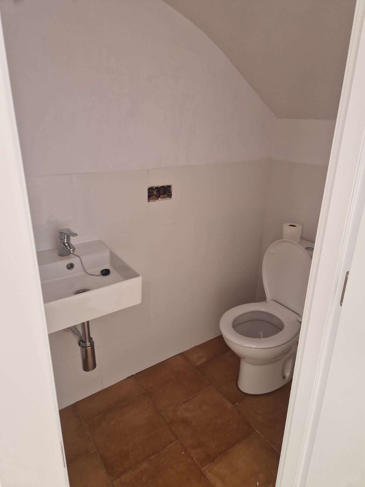 Baño pequeño, blanco, con inodoro y lavabo, suelo marrón, paredes blancas y techo empotrado.