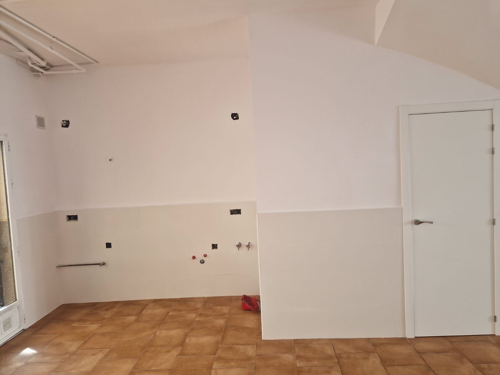 Habitación vacía con suelo de baldosas, paredes blancas, puerta y cableado expuesto.