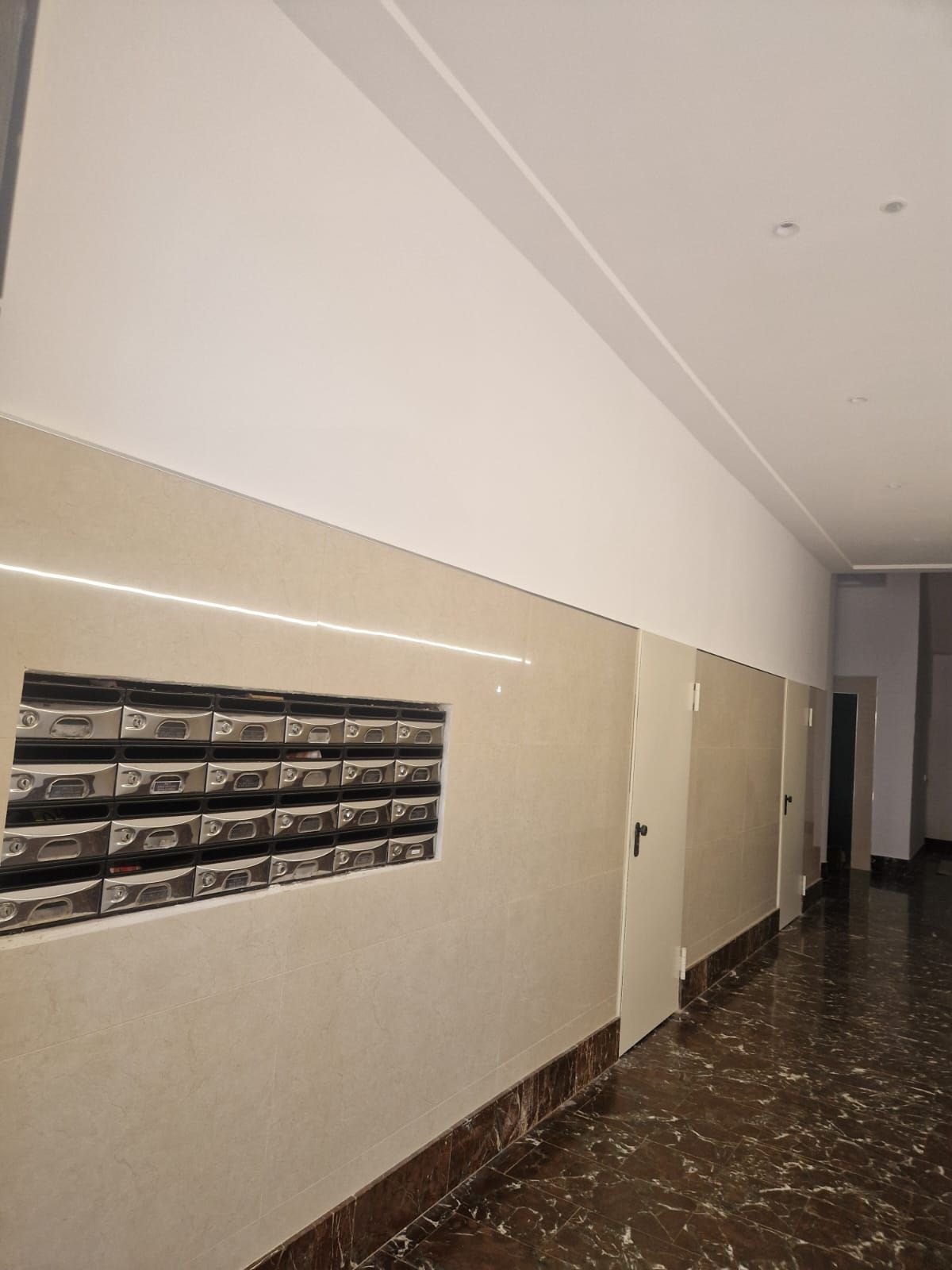 Pasillo con buzones, paredes beige, suelo y puertas oscuros.