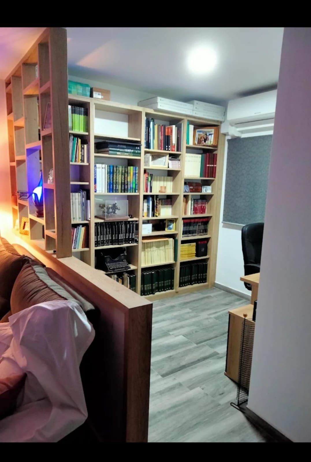 Estanterías que separan una sala de estar, llenas de libros y diversos artículos. Estantería beige, suelo gris.