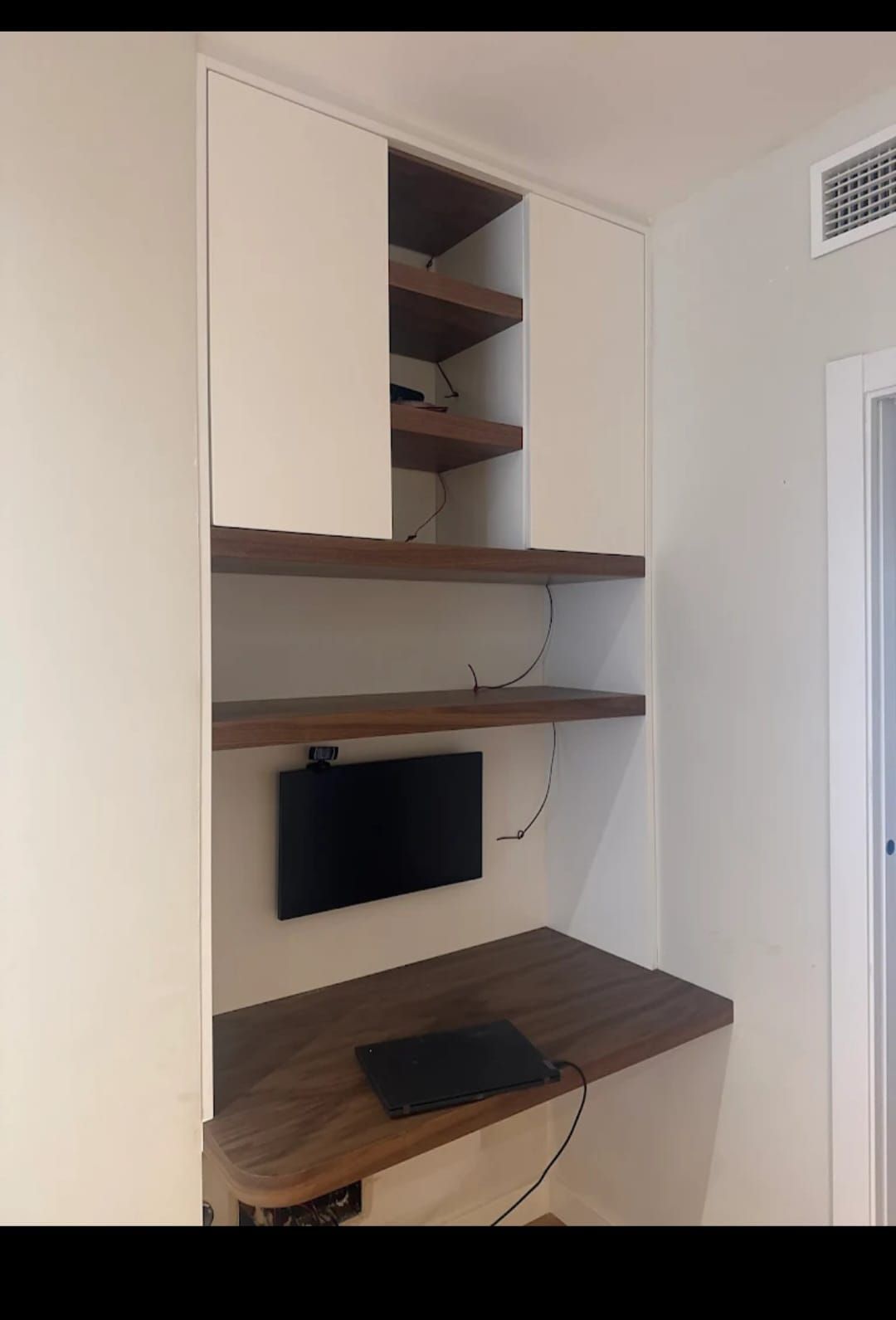 Escritorio empotrado con estantes de madera y puertas blancas en un nicho de pared. Sobre él hay una laptop y un monitor.