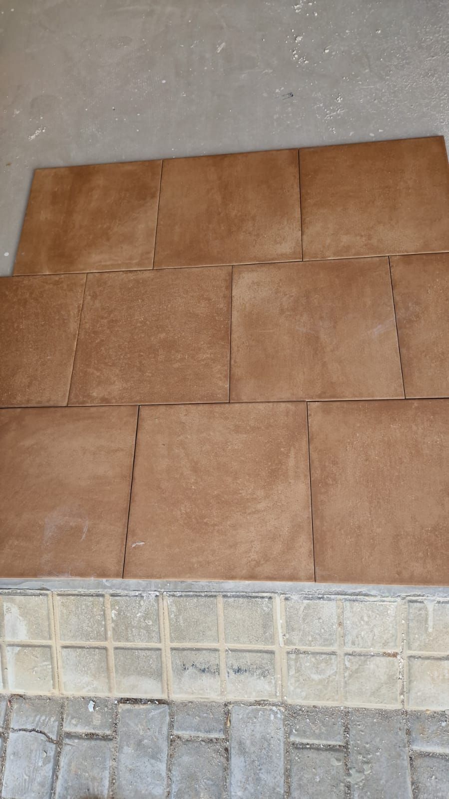 Azulejos marrones dispuestos sobre una superficie de hormigón, probablemente para su instalación, con el borde de un ladrillo decorativo en la parte inferior.