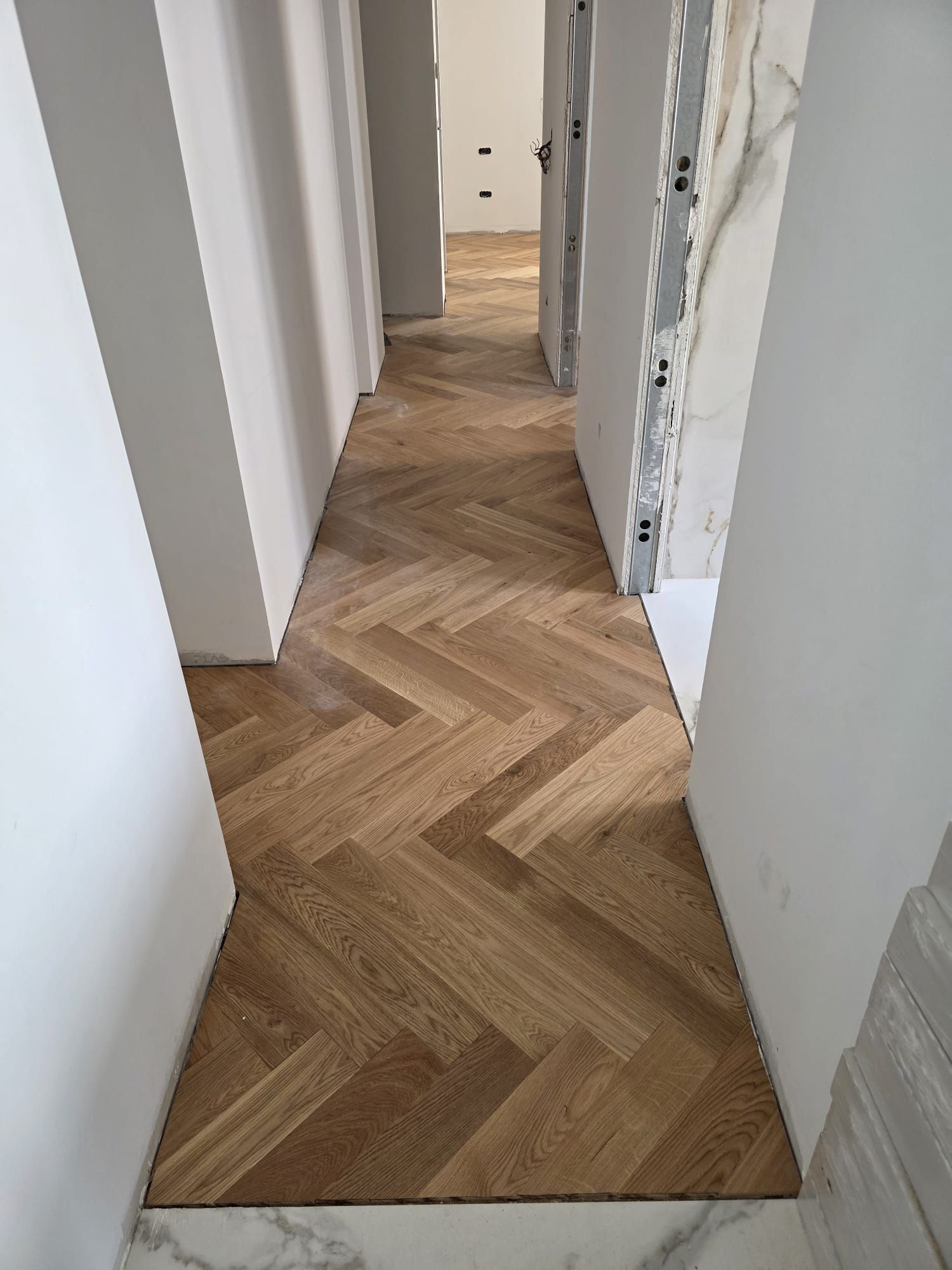 Piso de madera en espiga en un pasillo estrecho, rodeado de paredes blancas y puertas sin terminar.